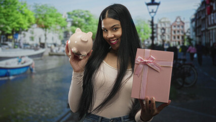 Woman holding pink piggybank and pastel giftbox on busy city street beside canal; financial planning optimism.