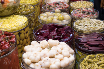 Wide assortment of pickled vegetables packed in brine inside glass jars at a market stall, rich color and texture; for food design, retail signage, recipe content, education.