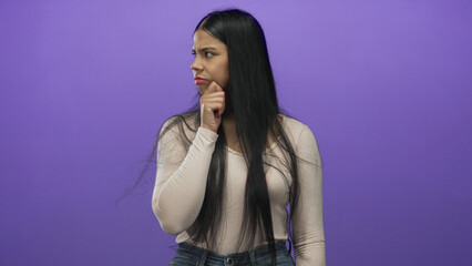 Woman with hand on chin against purple studio backdrop with focused gaze and long black hair...