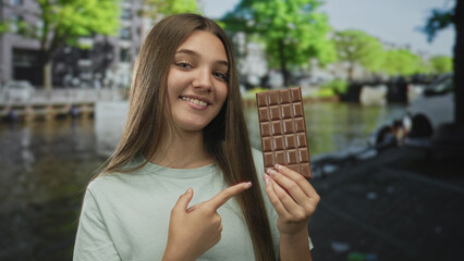 Fototapeta premium Smiling girl holds chocolate bar near canal water outdoors in amsterdam; delight indulgence sweet moment.