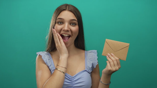 Teenage girl holds envelope with her right hand and touches cheek while smiling at camera in a teal studio; excitement.