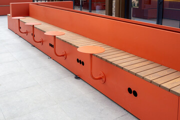 Long orange bench with integrated wooden seats and round tables on metal base at facade of modern building. Use for architecture, public spaces, urban furniture, landscape design. Real photo.
