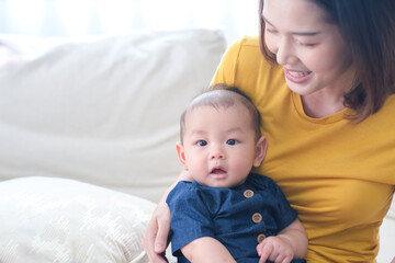 Loving asian mom carying of her newborn baby at home. Mother and baby boy playing in sunny bedroom. Parent and little kid relaxing at home. Family having fun together. Childcare, maternity concept.
