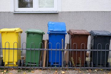 Row of colored recycling bins behind a fence outside a building, used for sorting waste.