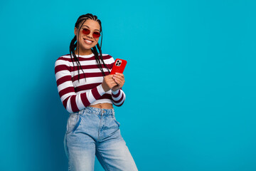 Naklejka na ściany i meble Young woman with striped top and jeans smiles while holding a red smartphone against a blue background Naklejka na ściany i meble Young woman with striped top and jeans smiles while holding a red smartphone against a blue background