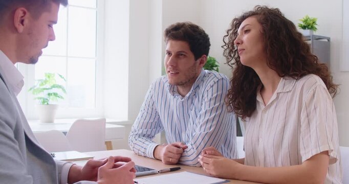 Discussion between couple and real estate agent about agreement document during consultation at office. People discuss property details, fees and signing steps with broker, sitting at desk at meeting