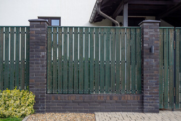Dark green wooden fence with brick pillars and gravel base near residential house. For showcasing privacy fencing, property boundaries, and modern front yard entrance design. Real photo