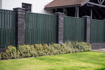 Green wooden fence with brick pillars and variegated hedge along neat lawn. For showcasing property borders, front yard landscaping, and low-maintenance garden edge design. Real photo.