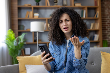 Young woman with curly hair feeling frustrated and confused, holding a smartphone and gesturing...