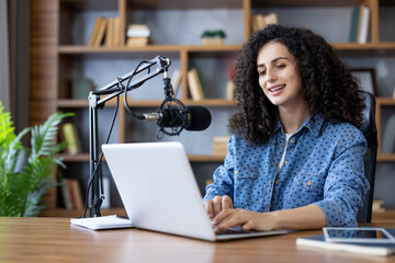 Woman content creator recording a podcast and typing on a laptop with a professional microphone in a home studio, creating digital content for her audience