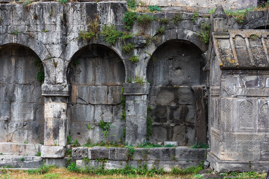 View of weathered gray stone arches and a crypt stand against a backdrop of an ancient stone wall, accented by patches of green grass, Tatev, Syunik Province, Armenia.