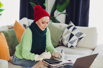 Elderly woman in cozy winter attire using a calculator at home office on laptop for finances