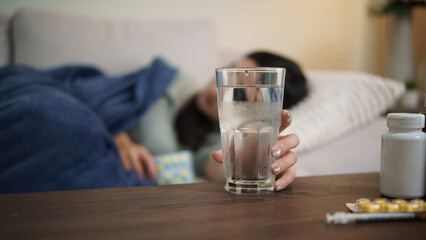 Sick woman lying on sofa reaching for a glass of water, feeling weak and tired while recovering from illness at home.