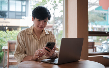 Fototapeta premium Smiling young man interacting with his mobile phone while working on laptop in a modern cafe.