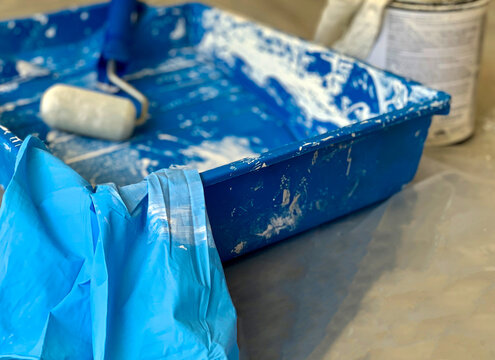 Low angle view of painting tools during a painting project with blue paint tray and white paint on it, a small painting roller, blue latex gloves and partial view of painting can in background. 