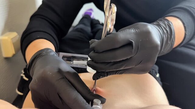 Macro close-up of piercer performing belly button piercing using sterile tools and needle. Focus on hands, jewelry, and professional technique. Clean, hygienic salon environment.