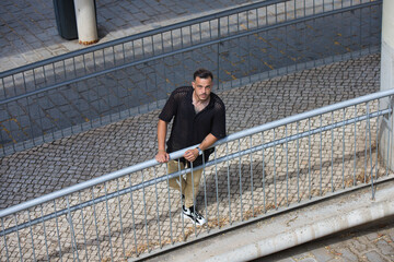 A young, handsome man with a beard, wearing a black shirt and beige cargo trousers, is holding onto...