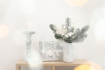Beautiful composition with fir tree branches in vase, gift and candles on wooden table near white wall, bokeh effect. Winter decor