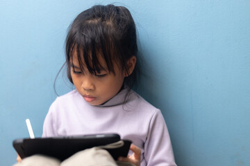 Asian little girl using tablet with stylus and focusing on study on blue wall background, concept of digital education, smart learning, e-learning, and technology for kids.