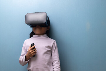 Asian little girl wearing VR headset and holding controller for learning and digital experience on blue wall background, concept of virtual reality, metaverse, and future education technology.