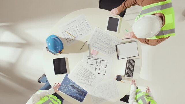 Top angle of construction professionals reviewing architectural drawings on round table. Concept of precision, teamwork, and modern project planning in engineering collaboration.