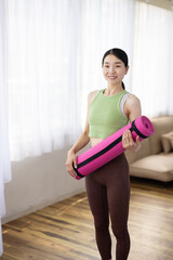 Smiling Asian Middle-Aged Woman in Activewear Holding Yoga Mat Indoors