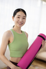 Smiling Asian Middle-Aged Woman in Activewear Holding Yoga Mat Indoors