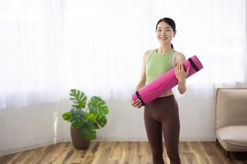Smiling Asian Middle-Aged Woman in Activewear Holding Yoga Mat Indoors