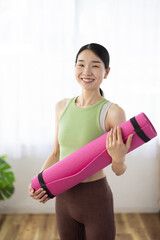 Smiling Asian Middle-Aged Woman in Activewear Holding Yoga Mat Indoors