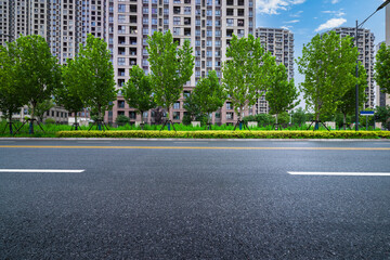 Empty asphalt street with a row of green trees in front of modern residential apartment buildings.