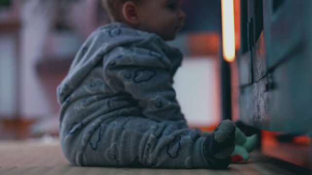 Baby sitting on floor beside wooden armoire, leaning slightly forward and looking curiously inside, dressed in soft grey pajamas with cloud pattern, evoking early exploration