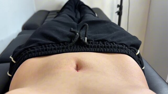 A close-up of a female navel piercing procedure. A gloved hand holds a piercing tool near the belly button. The background is blurred, focusing on the action.