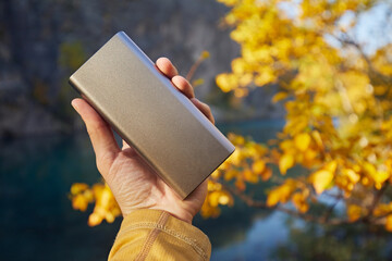 A man's hand holds a power bank with a charging phone against the backdrop of an autumn landscape