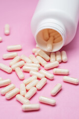 White capsules or tablets poured out of plastic bottle on pink background.