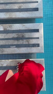 A young woman in a red dress walks across a bridge over a swimming pool. A view of her beautiful legs.