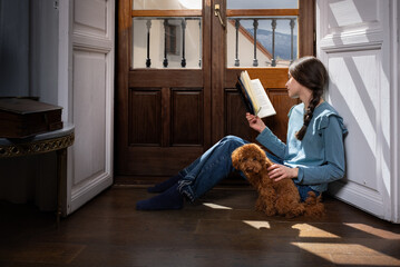 Preteen girl reading a book by the window with a brown poodle by her side. Cozy indoor moment showing childhood, pet companionship and peaceful home lifestyle.