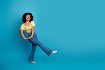 Joyful young woman in yellow top posing casually with vibrant blue background in trendy jeans and sneakers