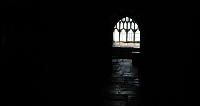 A lone figure in silhouette walks past an arched gothic window inside a dim medieval church corridor. Deep shadow and stone floor create a mood of solitude, reflection, and timeless faith.