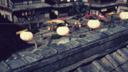 Soft lantern light glows in the evening as people enjoy the peaceful atmosphere of a traditional courtyard in a historic village. The rooftops frame the charming view below, inviting serenity.