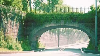 A peaceful scene features a stone archway framed by vibrant foliage. The sun shines brightly, casting soft shadows on the winding road beneath, creating a calm atmosphere.
