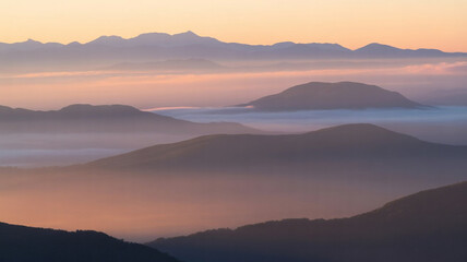 Fototapeta premium Serene mountain peaks shrouded in soft morning mist under a warm golden sunrise sky