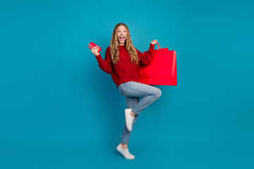 Charming young woman in casual red sweater holding shopping bag and smartphone against blue...