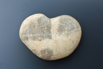 Close-up of a smooth, heart-shaped river stone with a natural dark cross pattern, isolated on a dark gray background. Symbolizes love, nature, meditation, healing, and abstract texture.