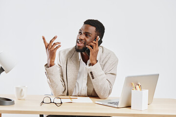 Smiling young African American man talking on smartphone while working at desk in modern office setting, using laptop for communication and multitasking, casual fashion, positive mood, studio shot