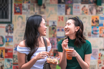 Two friends happily eating street snacks and drinking iced beverages outdoors, showing asian lifestyle, casual social interaction, friendly atmosphere, joyful candid expressions and tasty food culture