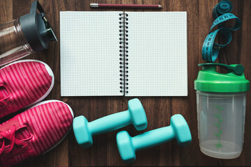 Top view of open notebook with pencil, pink sneakers, blue dumbbells, shaker, water bottle and measuring tape on wooden background with copy space, fitness planning and workout equipment flat lay