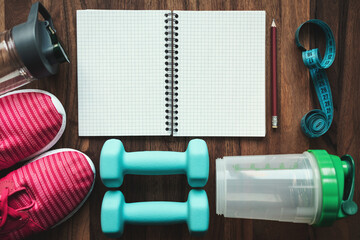 Top view of open notebook with pencil, pink sneakers, blue dumbbells, shaker, water bottle and measuring tape on wooden background with copy space, fitness planning and workout equipment flat lay