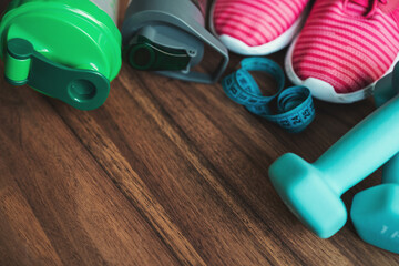 Blue dumbbells, pink sneakers, water bottle, protein shaker and measuring tape on wooden background with copy space, fitness equipment concept