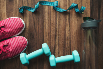 Top view of pink sneakers, blue dumbbells, water bottle and measuring tape on wooden background with copy space, fitness equipment flat lay concept