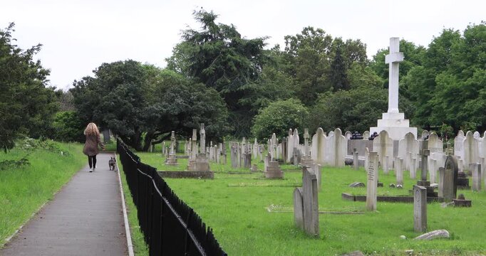 A woman with blonde hair walks a small dog along a path beside rows of historic headstones and a large memorial cross in a peaceful green cemetery, suggesting memory, time, and everyday life near deat